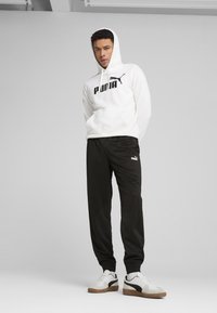 Hanorac alb cu logo negru "PUMA", asortat cu pantaloni de trening negri ce au un mic logo PUMA. Încălțăminte: adidași PUMA albi și gri cu accente maro.