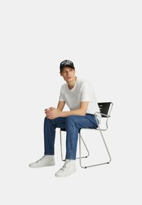 T-shirt bianca, jeans blu, sneakers bianchi e berretto nero con "MILANO 1984" in bianco. Seduto su una sedia nera con struttura cromata, sfondo neutro.