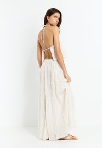 Lichtbeige maxi-jurk met open rug, halternek met strik en gathered taille; heeft vloeiende stof met subtiele sequindetails.