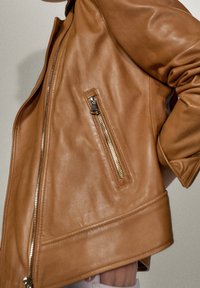 Giacca in pelle marrone con texture liscia, dotata di una tasca con zip sul lato. Il design presenta una vestibilità rilassata e linee pulite.