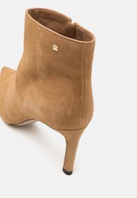 Bottine en daim camel avec talon haut et fin, fermeture éclair arrière, et petite plaque logo carrée dorée près du bord supérieur.