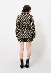 Patroon outfit in beige en zwart bestaat uit een blouse met kraag en bijpassende shorts. Het materiaal lijkt textuur te hebben; de blouse is op de taille geknoopt. Zwarte enkelschoenen.