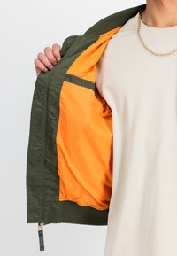 Maslinasto zelena jakna s rebrastim ovratnikom, s narančastom podstavom. Unutrašnjost je glatka, s patentnim zatvaračem na rubu.