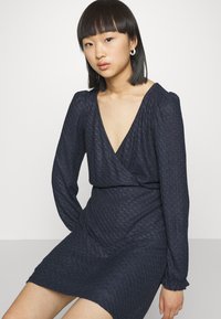 Vestido envolvente azul marinho com mangas longas bufantes, decote em V profundo, tecido texturizado e cintura ajustada, alcançando a metade da coxa. Brincos em forma de argola.