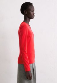 Czerwona bluzka na długi rękaw z okrągłym dekoltem, wykonana z gładkiej tkaniny, zestawiona z szarą spódnicą z plisami, widziana z boku.