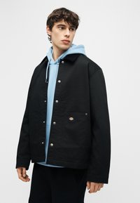 Νέος άνδρας που κοιτάζει πλάγια, φοράει μαύρο μπουφάν με κουμπιά πάνω από ένα ανοιχτό μπλε φούτερ με κουκούλα, σε απλό φόντο.