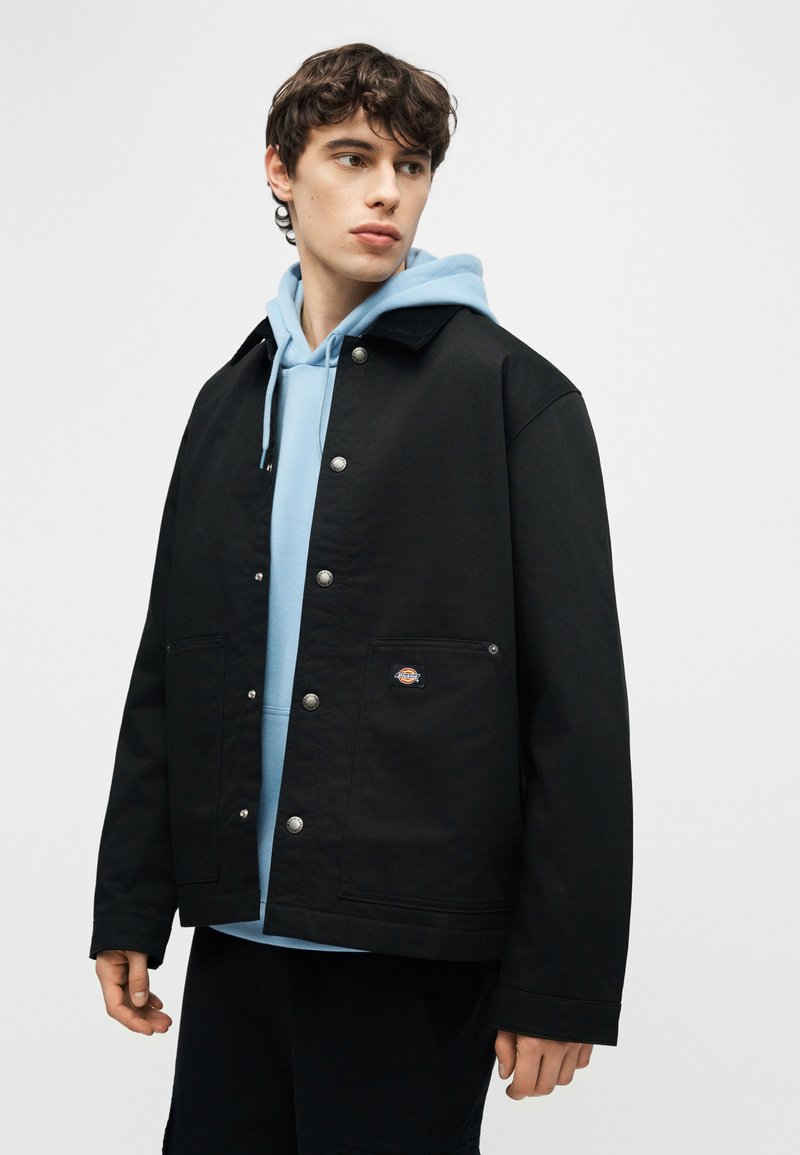 Giovane uomo che guarda di lato indossando una giacca nera con bottoni sopra una felpa con cappuccio azzurro chiaro, su uno sfondo neutro.