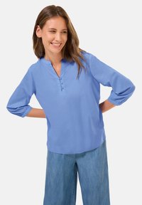 Blusa azzurra chiara con scollatura a V, tre bottoni frontali e maniche a tre quarti, realizzata in un tessuto morbido e con una vestibilità rilassata.