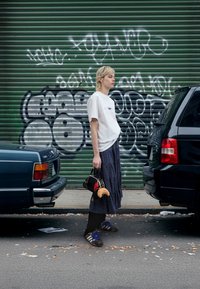 Hvit t-skjorte, langt marineblått rutete skjørt, brune joggesko med blå detaljer. Svart håndveske med rød blomst og koseleke. Urban graffiti-bakgrunn.