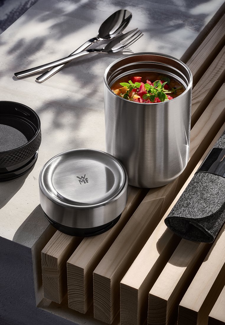 Pojemnik na jedzenie ze stali nierdzewnej z zupą i ziołami, pasującą pokrywką, metalową łyżką i widelcem oraz szarym materiałowym pokrowcem na sztućce na drewnianych listwach.