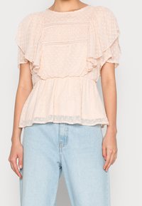 Blouse rose clair avec un motif à pois, manches à volants, et un détail de smocks texturé au niveau du buste, taille peplum et ourlet transparent.