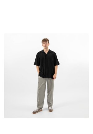 Mladý muž stojící s rukama v kapse, oblečený v černé košili na knoflíky s krátkým rukávem, šedých kalhotách a šedých teniskách na bílém pozadí.