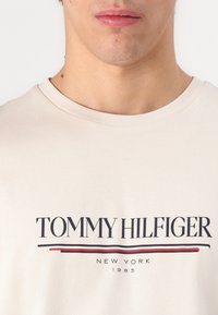 Beige pamut póló, amelyen a vastag fekete "TOMMY HILFIGER" felirat szerepel, alatta piros és kék csíkkal, könnyű anyagú, klasszikus kerek nyakkivágással.