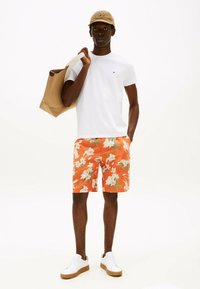 Camiseta blanca, pantalones cortos florales naranjas, zapatillas blancas y una gorra beige con un logo. Llevando una bolsa de tote marrón. Atuendo sencillo y casual de verano.