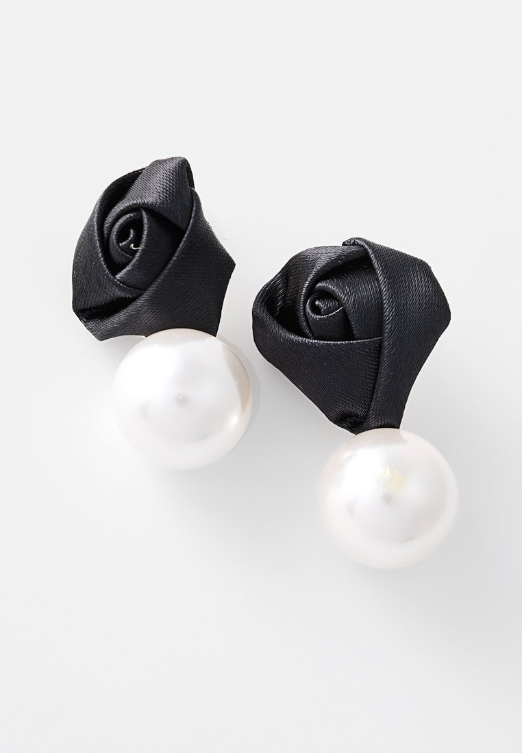 Pendientes de diseño en satén negro con flores que presentan grandes perlas blancas. Los pétalos están intrincadamente moldeados, contrastando con la superficie lisa de la perla.
