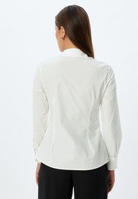 White fitted shirt with a subtle sheen, long sleeves, and a notched collar. Features pleating at the back and buttoned cuffs.