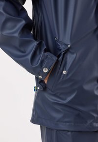 Marineblauwe waterdichte jas met een glanzende, gladde textuur; voorzien van drukknopen en een klein Zweeds vlagdetail aan de zoom.