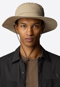 Sombrero beige de ala ancha con textura y correa ajustable negra en la barbilla, llevado con una camisa oscura sobre una blusa marrón.