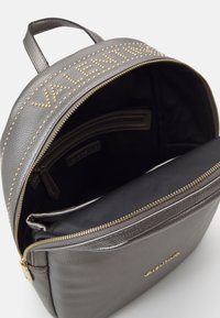 Grijze leren rugzak met studded letters aan de voorkant, met een ritssluiting hoofdcompartiment en een gladde, gestructureerde afwerking.