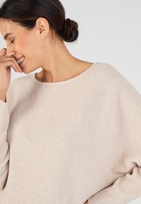 Maglione leggermente beige con texture a costine e scollo ampio. Caratterizzato da maniche morbide e un materiale soffice e accogliente.