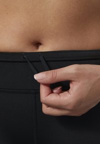 Zwarte sportlegging met een gladde textuur. De hoge tailleband heeft een trekkoord en de hand trekt aan de tailleband.