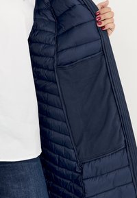 Marinblå puffy-jacka i quiltat tyg, med en slät innerfoder och en inre ficka. Utsidan har en texturerad och isolerad design.