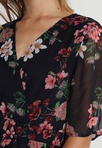 Robe fleurie en tissu noir transparent, ornée de fleurs roses et blanches, avec un décolleté en V et des manches courtes à la coupe fluide.