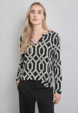 Top a maniche lunghe con motivo geometrico in bianco e nero, con scollatura a V e tessuto leggero, abbinato a pantaloni neri.
