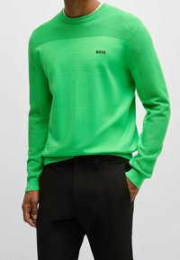 Grön stickad tröja med ribbat mönster på axlarna, med rund halsringning och liten svart "BOSS"-logotyp. Bärs med svarta byxor.