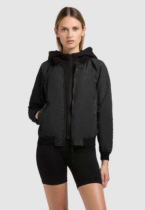 Ung kvinde iført en sort hættetrøje med lynlås og sorte cykelshorts, stående med den ene hånd i lommen, neutral baggrund.