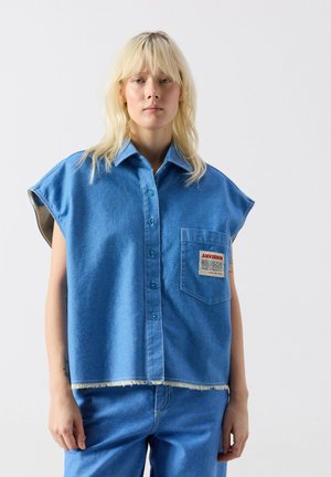 Blonde person wearing sleeveless blue denim button-up shirt with front pocket and matching blue denim pants, standing against plain background.