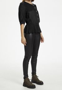 Blusa negra texturizada con mangas abullonadas, escote redondo y botones frontales. Combinada con pantalones ajustados de imitación cuero negros y botas oscuras con cordones.