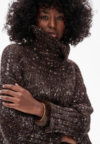 Kobieta z kręconymi włosami, ubrana w brązowy sweter z grubym splotem i golfem, z założonymi na siebie rękami i złotą bransoletką na białym tle.