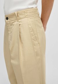 Beige trousers with a flat front and pleats, made of lightweight fabric. Features a side pocket and a label on the waistband.