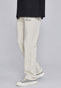 Bežā krāsā sporta bikses, izgatavotas no mīksta auduma, ar izšūtu logotipu kreisajā augšstilbā, kopā ar pelēkiem un baltiem sporta apaviem. Modelis valkā melnu krekliņu.