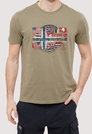 Homme portant un t-shirt vert olive avec un imprimé style vintage de plusieurs drapeaux internationaux, associé à un short cargo foncé.