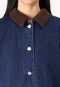 Jeanstjacka i mörk indigo med krage i brunt cordtyg, prydd med silverfärgade tryckknappar och kontrasterande sömmar framåt.
