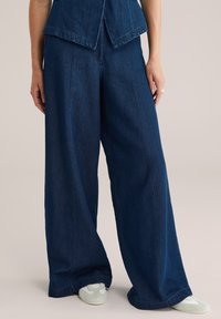 Platām kājām izgatavotas tumši zilas denima bikses, ar gludu tekstūru, augstu jostasvietu un ikdienišķu piegriezumu. Apvienotas ar gaišas krāsas apaviem.