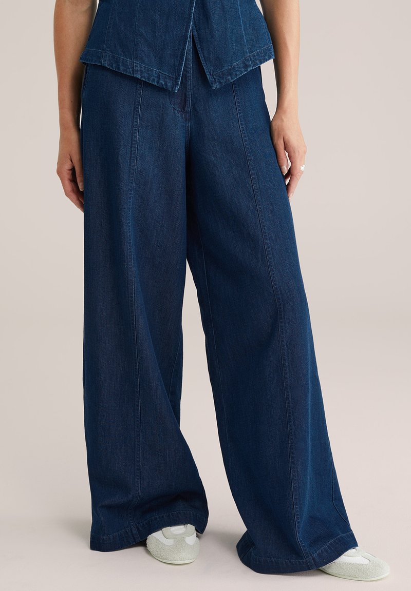 Platām kājām izgatavotas tumši zilas denima bikses, ar gludu tekstūru, augstu jostasvietu un ikdieni�šķu piegriezumu. Apvienotas ar gaišas krāsas apaviem.