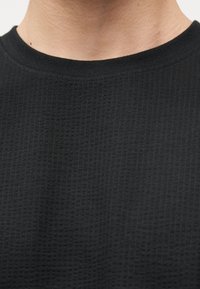 Κοντινό πλάνο σελαιμού και άνω μέρους στήθους ενός ατόμου που φοράει μαύρο, υφασμάτινο μακρυμάνικο πουλόβερ με υφή βάφλας και στρογγυλή λαιμόκοψη.