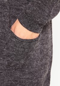 Grå stickad tröja med synlig ficka och mudd. Tyget ser mjukt och fluffigt ut, med en blandad, melerad look.