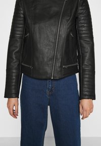Giubbotto da moto in pelle nera con accenti a coste sulle spalle, due tasche con cerniera e cerniera frontale asimmetrica. Abbinato a jeans in denim blu.