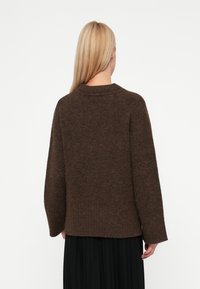 Pulôver castanho de malha com decote arredondado, ombros caídos e um acabamento texturizado. A parte de trás é ligeiramente mais longa do que a frente.