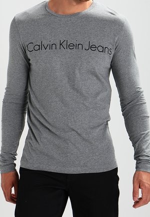 Mężczyzna w dopasowanej szarej koszuli z długim rękawem z napisem "Calvin Klein Jeans" na piersi i czarnych spodniach.