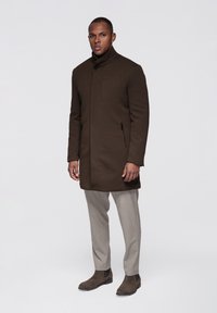 Tmavě hnědý kabát s vysokým límcem, dvěma bočními zipovými kapsami a úzkým střihem. Nošený přes světlé šedé kalhoty a hnědé chelsea boty.