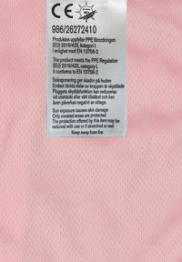 Rosa tygetikett med vit text. Innehåller skötselråd och efterlevnadsinformation för personlig skyddsutrustning (PPE) enligt EU-föreskrifter.