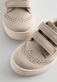 Beige shoes with Velcro straps, textured fabric upper, scalloped edge design, and perforated detailing on the toe. Rubber outsole.