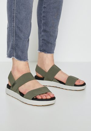 Fødder iført oliengrønne stofremssandaler med tykke hvide såler, parret med blå jeans med frynset kant mod en ensfarvet lys baggrund.