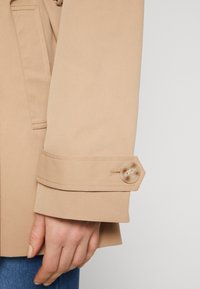 Beige, gestructureerde stoffen jacksleeve met een hexagoonvormige manchette, voorzien van een grote, lichtgekleurde knop en zichtbare stikdetails.