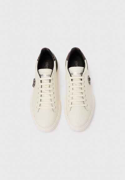 Ett par vita lädersneakers med snörning, metalliskt sidologotyp, mörk inredning och mönstrad häldetalj, vy ovanifrån på vit bakgrund.